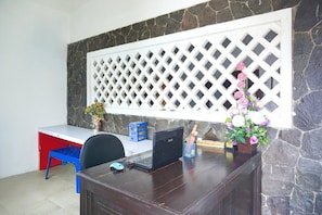 A wooden desk with a laptop, a blue chair, a decorative vase with flowers, and a stone wall.