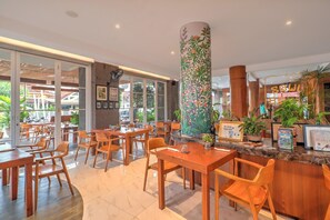 A restaurant with wooden tables and chairs, a large decorative pillar, and a counter with plants and framed items.