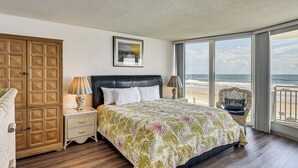 Een slaapkamer met een bed, nachtkastjes, een houten garderobe, uitzicht op het strand en een rieten stoel.