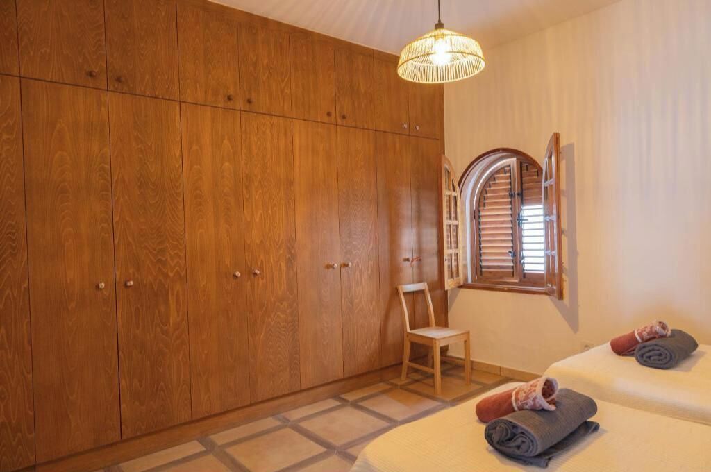 A bedroom with a large wooden wardrobe, a chair, a bed with towels, and a hanging light fixture.