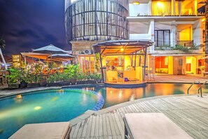 Zona de piscina con cubierta de madera, área de descanso con sombrillas y un edificio con estructura cilíndrica.