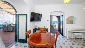 Un'area pranzo con tavolo e sedie in legno, una televisione montata a parete e vista mare attraverso un'apertura.