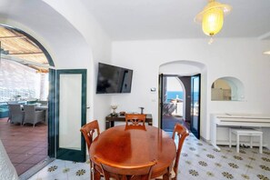 A dining area with a wooden table and chairs, a television mounted on the wall, and a view of the sea through an open door. - Clifftop Sea Views & Panoramic Terrace (Conca dei Marini)