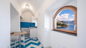 Une cuisine compacte avec un sol carrelé bleu et blanc, un coin repas avec des chaises blanches et une fenêtre donnant sur un paysage côtier.