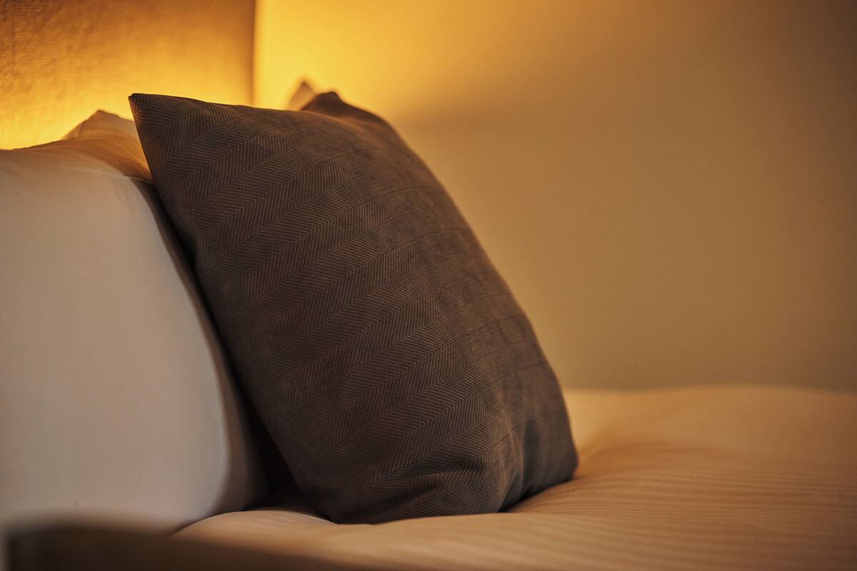 Ein Detailaufnahme eines Bettes mit Kissen, einer gemusterten Decke und einer Wand mit einer Lichtquelle.
