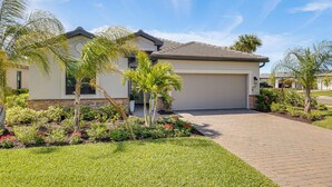 A residential house with a garage, a paved driveway, and landscaped garden with palm trees and red flowers. - New Home in Verdana Village w/ Resort Amenities! (Estero)
