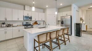 A modern kitchen with white cabinetry, a large island with bar stools, stainless steel appliances, and pendant lighting. - New Home in Verdana Village w/ Resort Amenities! (Estero)