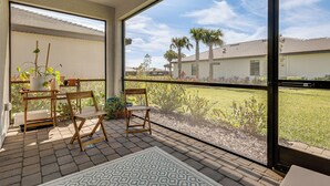 A balcony with a view of a grassy area and palm trees, a wooden chair, a small table with a plant, and a patterned rug. - New Home in Verdana Village w/ Resort Amenities! (Estero)