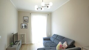 A living room with a grey sofa, a TV on a stand, a radiator, and a window with curtains. - Federica Flat - Metro Monte Grappa (Turin)