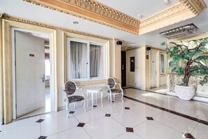 Un hall d’hôtel avec des détails ornementaux au plafond, une grande plante en pot et deux fauteuils près d’une fenêtre.
