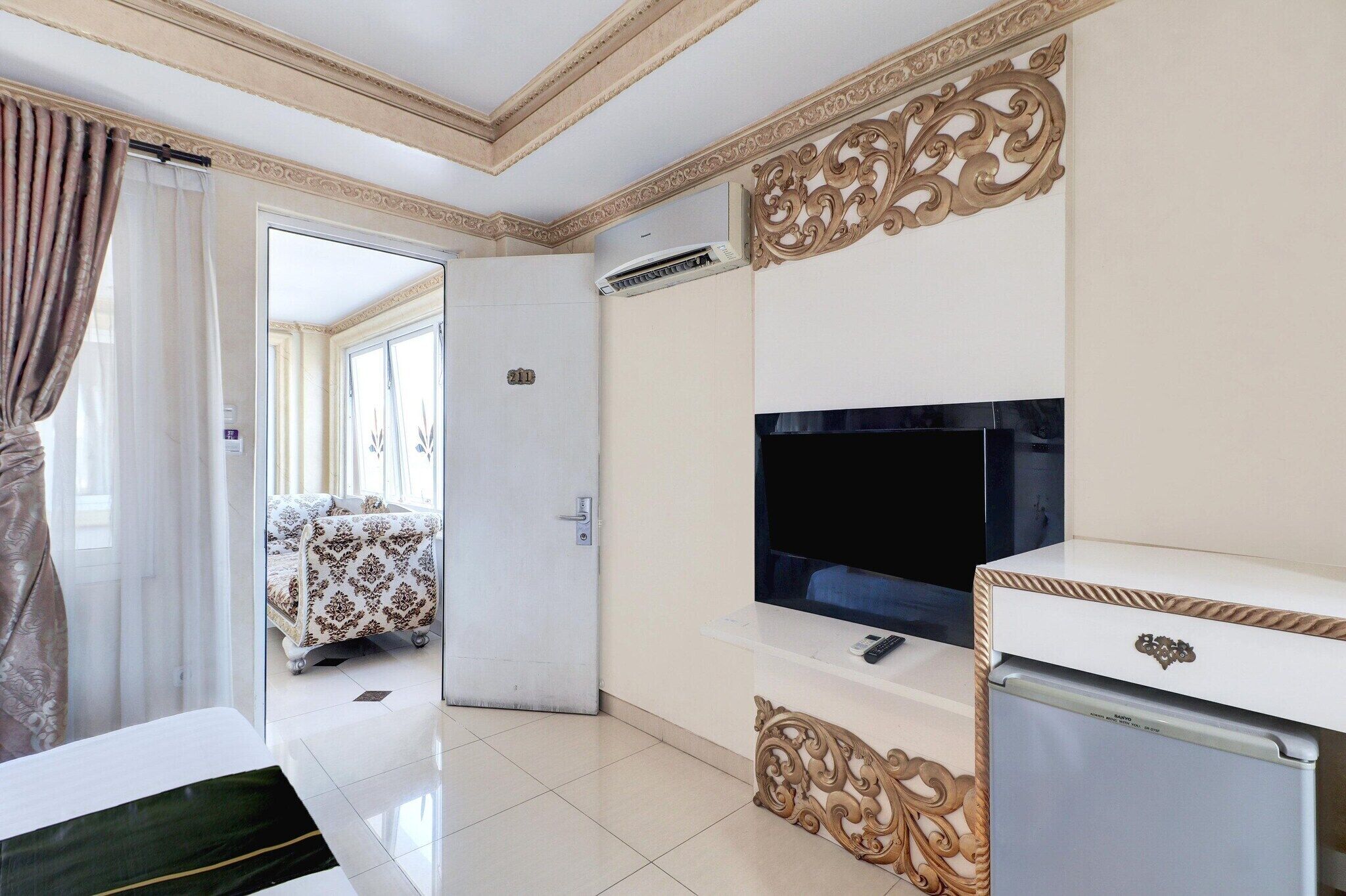 Habitación de hotel con televisión, refrigerador, cama con colcha estampada, puerta que da a otra habitación y un panel decorativo en la pared.