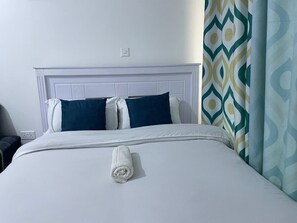 A neatly made bed with a white comforter and a rolled towel, a white headboard, and a patterned curtain. - Modern Cozy Studio-Next to TRM Mall (Nairobi)