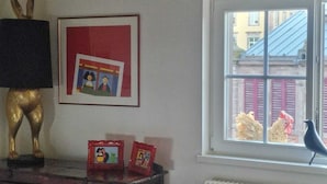 A room with a radiator, a window with a view of buildings, a framed picture on the wall, and a bird on the windowsill. - APARTMENT PANORAMIC VIEW OF PETITE VENISE PARKING (Colmar)