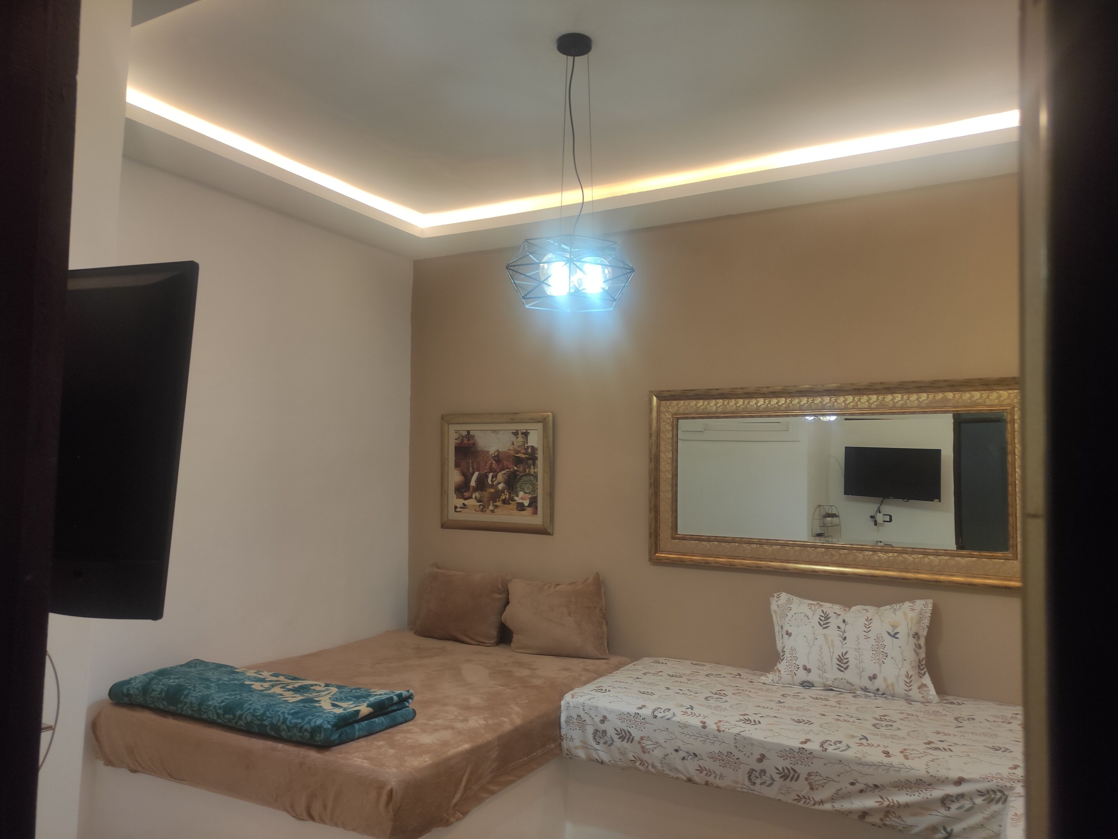 Un dormitorio con una cama, una cama adicional, una lámpara en el techo, un televisor montado en la pared y un cuadro enmarcado.