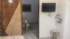 Una habitación con puerta de madera, un televisor montado en la pared, un taburete pequeño y una zona de comedor con sillas y una mesa.