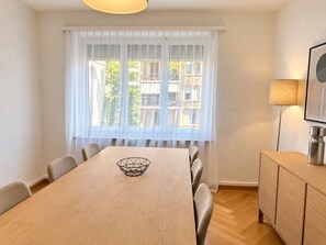 Ein Essbereich mit einem Holztisch, Stühlen, einer Dekoschale, einer Lampe und Blick durch ein Fenster mit durchsichtigen Vorhängen.