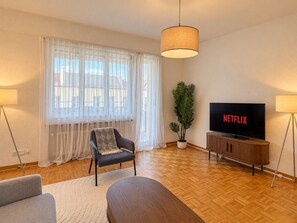 Ein Wohnzimmer mit einer Couch, einem Couchtisch, einem Fernseher auf einem Holzboden, einer Topfpflanze und durchsichtigen Vorhängen am Fenster.
