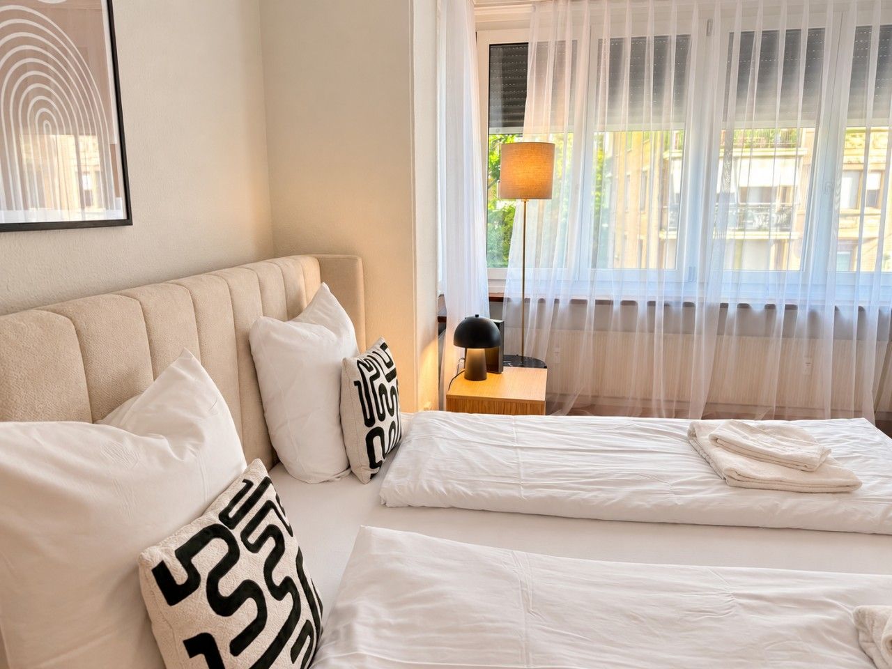 Et hotelværelse med seng, puder, et natbord, en lampe og et vindue med lette gardiner.