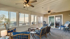 A covered outdoor seating area with wicker furniture, a ceiling fan, and large windows overlooking the ocean. - Gwynn's Island Gem | Private Beach & Bay Views (Hudgins)