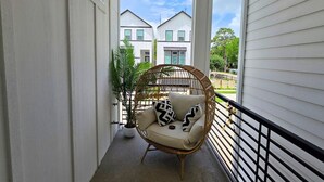 Een balkon met een rieten stoel, een bloempot en uitzicht op twee witte huizen.