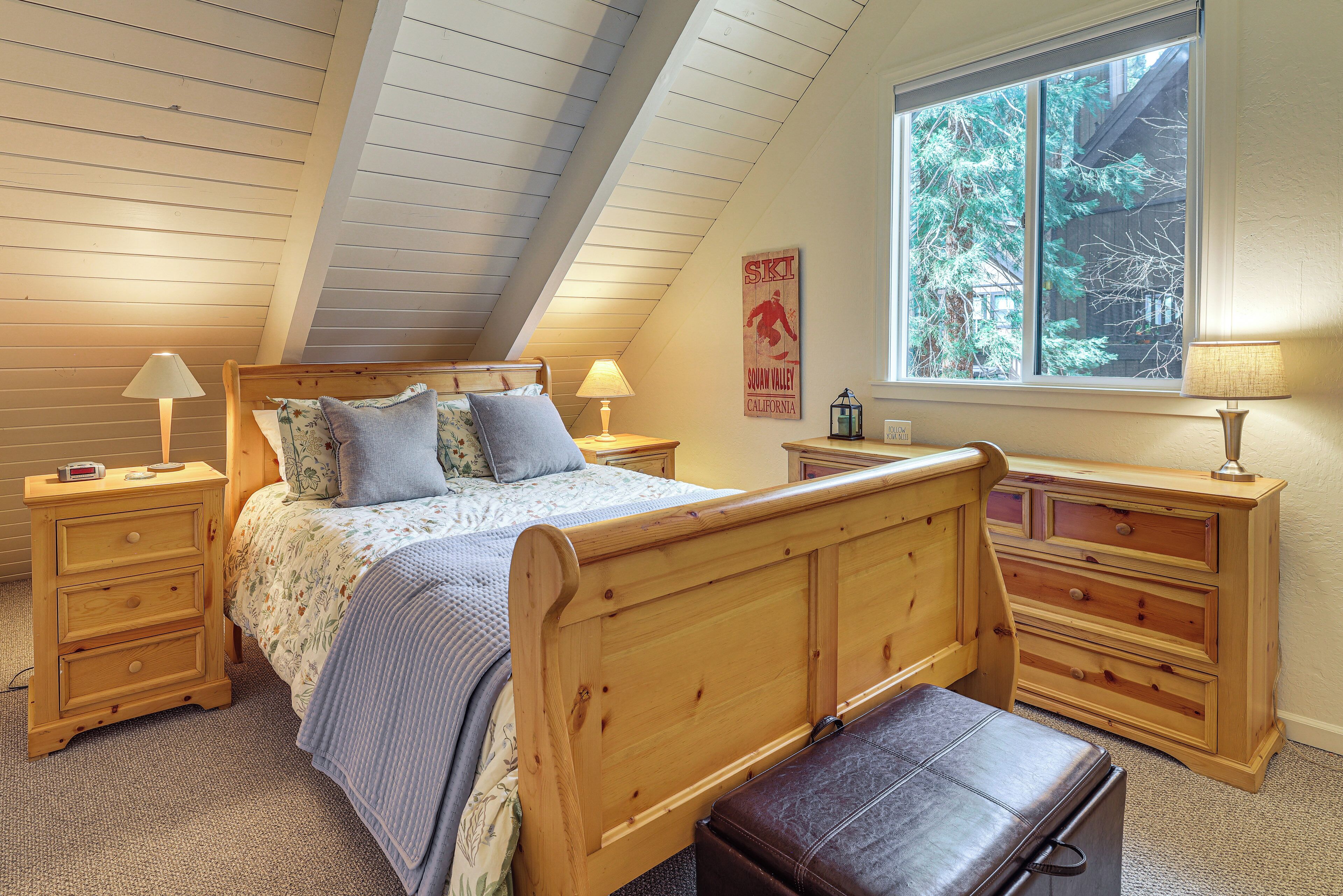 A bedroom with a sloped ceiling, a bed with a headboard, two nightstands, a dresser, and a window with a view of trees.