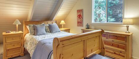 A bedroom with a sloped ceiling, a bed with a headboard, two nightstands, a dresser, and a window with a view of trees.