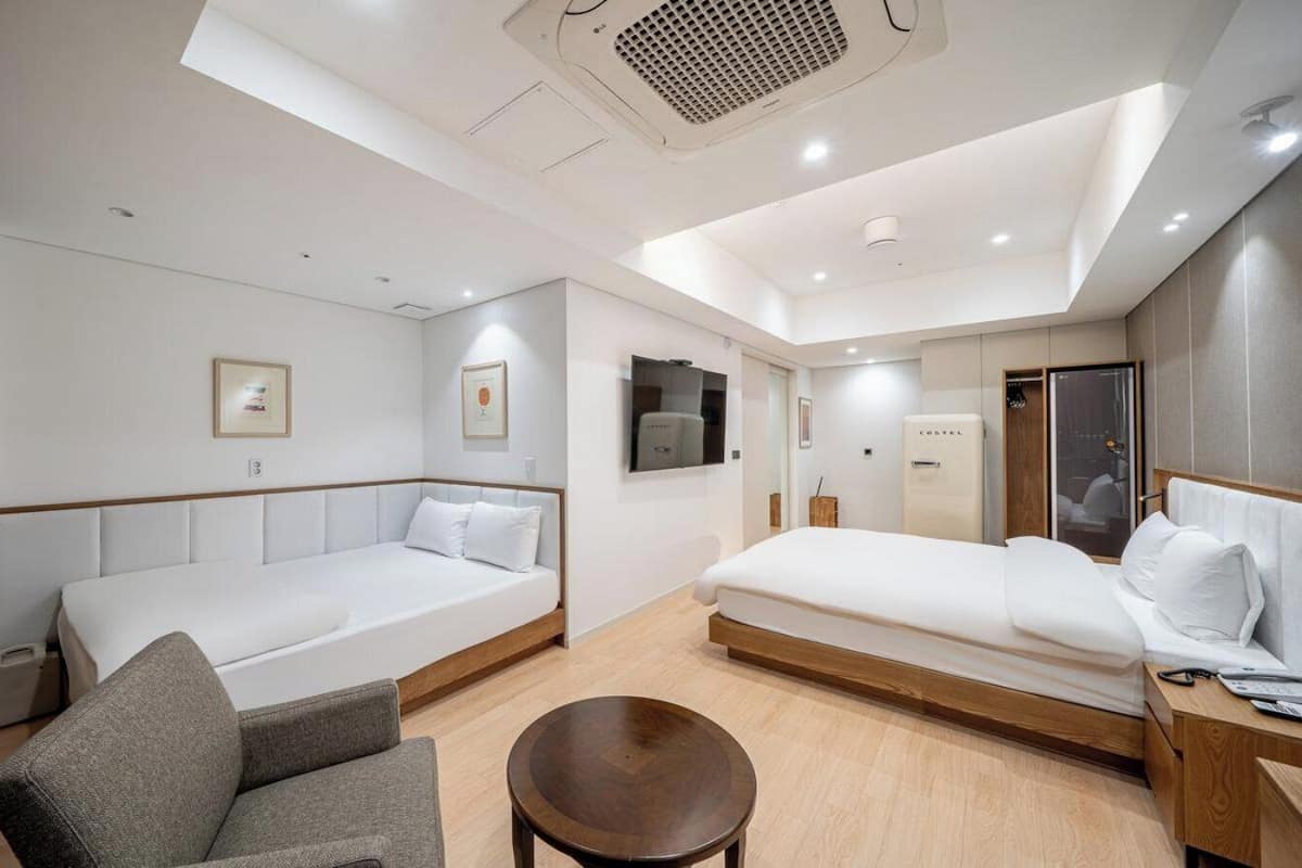 A modern hotel room with two beds, a sofa, a round table, and a shower area.
