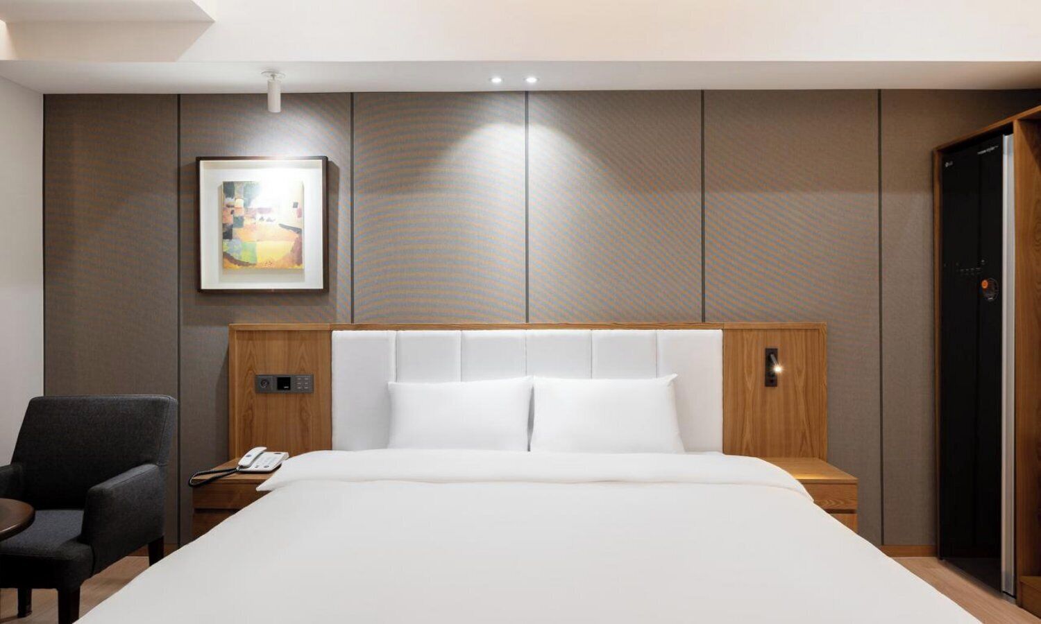 A hotel room with a large bed, a bedside table with a phone, a chair, a painting on the wall, and a wardrobe.
