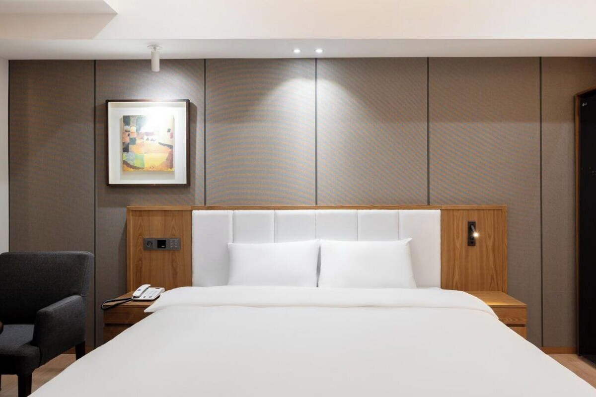 A hotel room with a large bed, a bedside table with a phone, a chair, a painting on the wall, and a wardrobe.