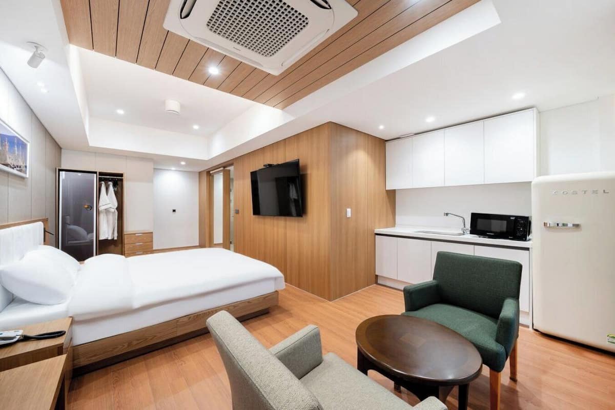A modern hotel room with a bed, seating area, kitchenette, and a television.