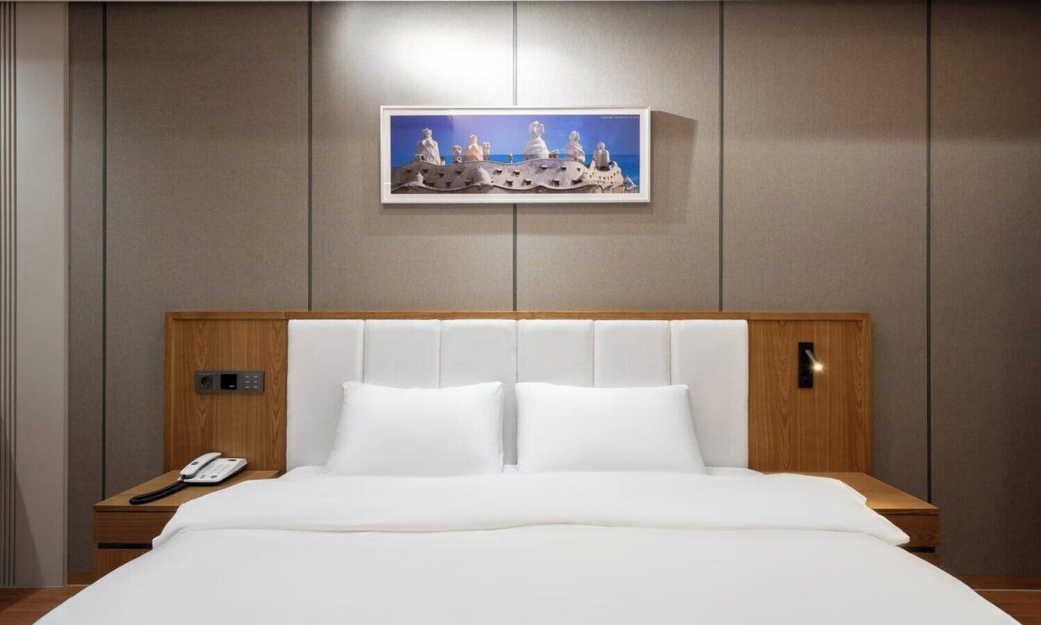 A hotel room with a large bed, a bedside phone, a framed artwork above the bed, and a wooden headboard.