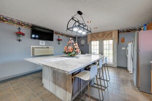 Una cocina con una isla de mármol, taburetes de bar, un refrigerador, un televisor y un arreglo floral decorativo.