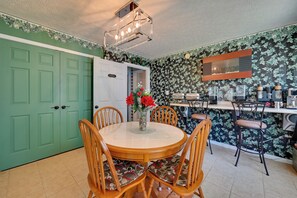 Un coin repas avec une table ronde, des chaises en bois, un vase avec des fleurs, un comptoir de cuisine avec des appareils électroménagers et une porte verte.