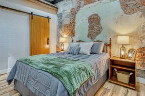 Une chambre avec un lit, une tête de lit en bois, deux tables de chevet avec des lampes, une commode en bois, un panier et un mur en briques apparentes.