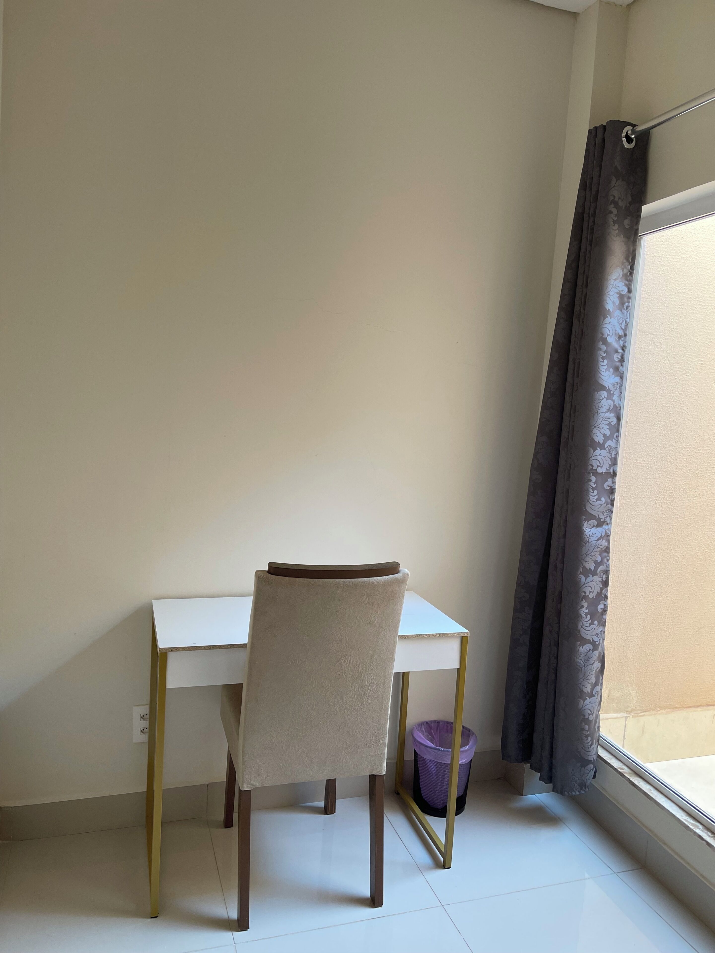 Une petite pièce individuelle, équipée d’un bureau et d’une chaise blancs, d’une fenêtre avec un rideau et d’une poubelle.