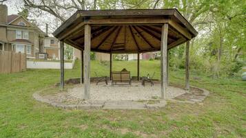A wooden gazebo with a bench and table, surrounded by a gravel area and grass, with houses in the background.