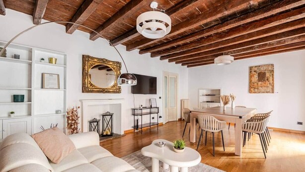 Una sala de estar con sofá, una mesa de comedor con sillas, chimenea, televisión y techo con vigas a la vista.