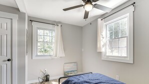 Un dormitorio con cama, ventilador de techo, dos ventanas con cortinas y un cuadro en la pared.
