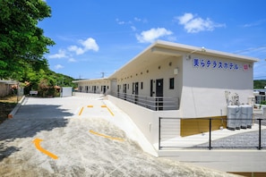 A hotel with a flat roof, multiple doors, and a fenced area with large water tanks. - Chura Terrace Hotel -AKI- (Nago)