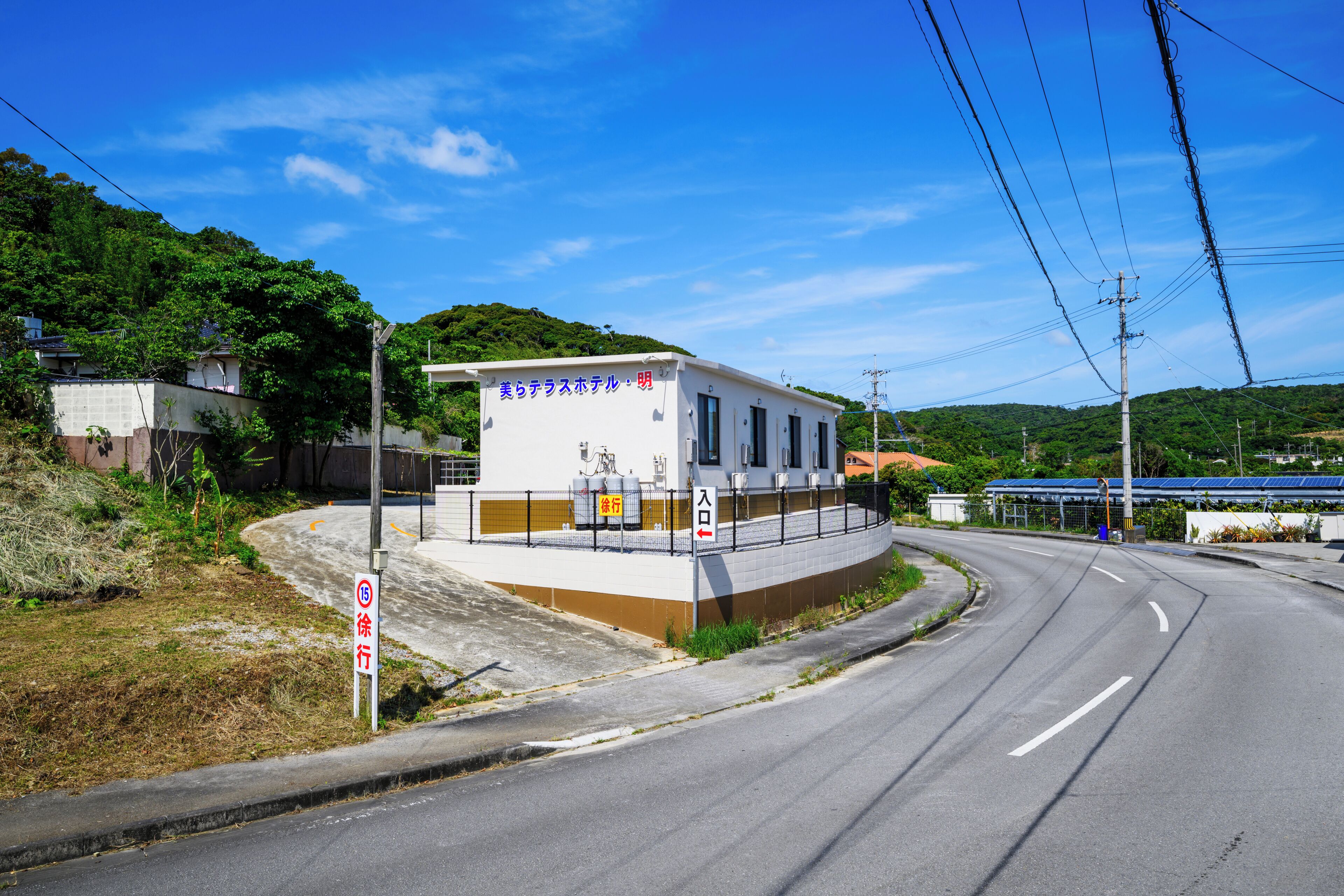 A small building with a sign in a foreign language, a road curving to the right, and a hill with greenery in the background.