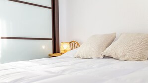 A bed with white bedding, two cream-colored pillows, a wicker chair, and a wall-mounted lamp. - Atlantic View Apartment - El Rompido (El Rompido)