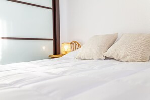 Un letto con lenzuola bianche, due cuscini color crema, una sedia in vimini e una lampada fissata al muro.
