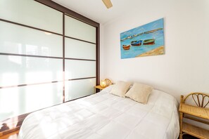 A bedroom with a bed, a nightstand, a lamp, a painting of boats on water, and a large window. - Atlantic View Apartment - El Rompido (El Rompido)