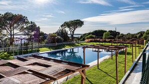 A pool with wooden loungers and a pergola, surrounded by a white fence and greenery, with a view of buildings and trees in the background. - Atlantic View Apartment - El Rompido (El Rompido)