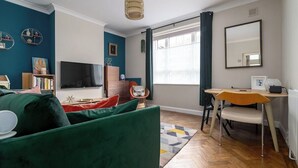 Olohuoneessa on vihreä sohva, televisio, puinen sivupöytä, tuoli ja ikkuna, jossa on verhot.
