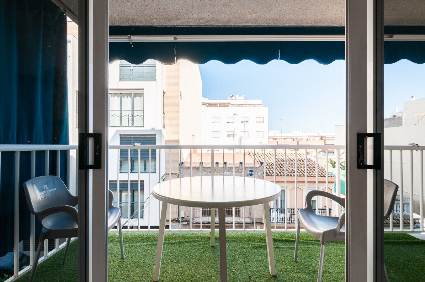 Een balkon met een tafel en stoelen, een uitzicht op gebouwen en een blauw zonnescherm.