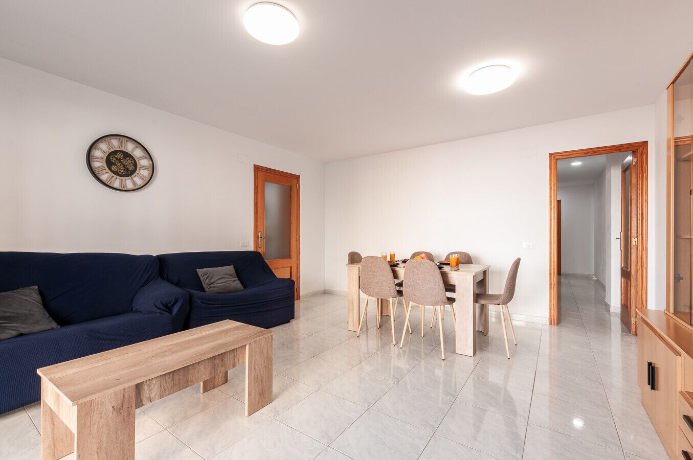 Una sala de estar moderna con un sofá, una mesa de comedor con sillas, un banco de madera y un reloj de pared.