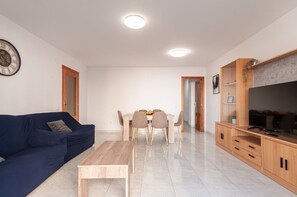 A modern living room with a blue sofa, a wooden coffee table, a dining area with a table and chairs, a TV on a wooden stand, and a clock on the wall. - Arenal Beach Escape - Burriana Port (Burriana)