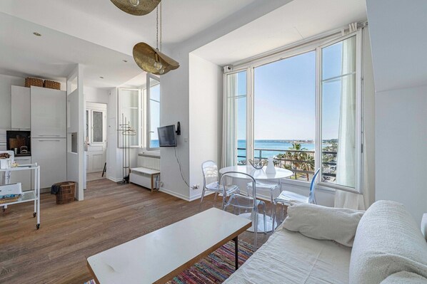 A modern, open-plan living space with a large window offering a view of the sea, a dining area, and a kitchen. - Air-conditioned Seafront Apartment – Nice (Nice)
