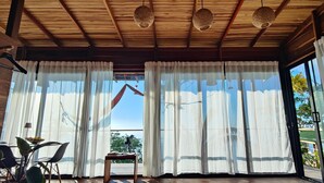 A room with wooden ceiling beams, white curtains, a table with a chair, and a view of the outdoors.